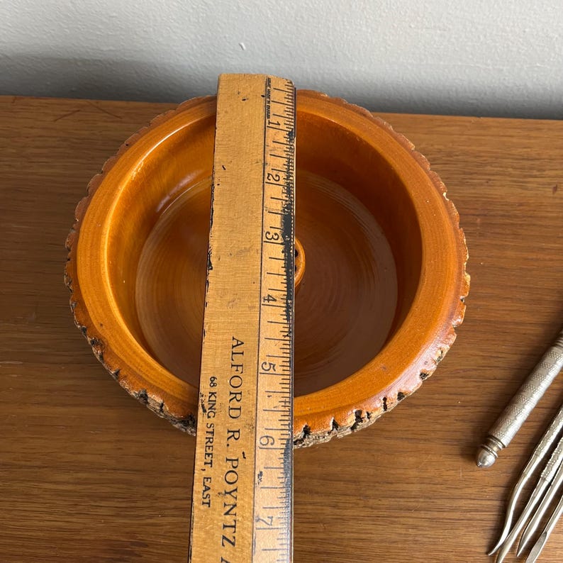May include: A wooden bowl with a natural bark edge, measuring approximately 6 inches in diameter. The bowl has a smooth, light brown interior and a rough, textured exterior. A vintage ruler is placed inside the bowl.