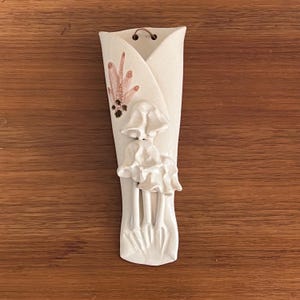 May include: Cream ceramic wall pocket vase with a floral and mushroom design. The vase has a curved shape and a small metal hanger. The design includes a cluster of mushrooms and a floral accent in shades of pink and brown.