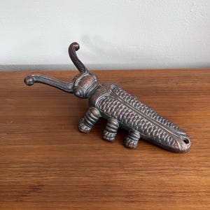 Może przedstawiać: Otwieracz do butelek z metalu w kolorze brązu w kształcie stylizowanego owada. Otwieracz ma długi, zakrzywiony uchwyt i szczegółowy korpus z teksturowanymi wzorami. Owad ma sześć nóg i zakrzywioną antenę.