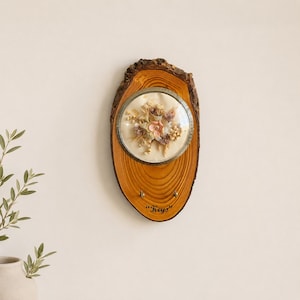 Op de afbeelding: Een houten sleutelhouder met een natuurlijke boomschorsrand. Het heeft een cirkelvormige weergave van gedroogde bloemen in een met glas bedekt frame. Het woord "Keys" staat onder de sleutelhaken.