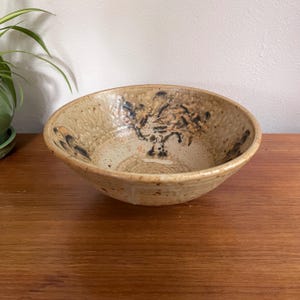 Peut inclure: Un bol en céramique fait à la main avec une finition mouchetée beige et marron. Le bol présente une surface texturée et un motif floral décoratif dans des tons marron et bleu. Le bol est posé sur une surface en bois.
