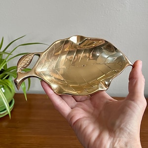 May include: A brass leaf-shaped dish with detailed veining. The dish is held in a hand, showcasing its polished, golden surface. The leaf design includes a stem and a detailed pattern, perfect for holding small items.