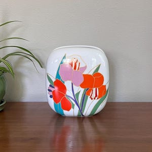 May include: White ceramic vase with a square shape and rounded corners. The vase is decorated with colorful floral designs in shades of red, orange, and pink, with green leaves and gold accents. The vase is sitting on a wooden surface.