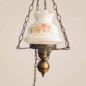 Peut inclure: Une lampe suspendue ancienne avec un abat-jour en verre blanc orné de motifs floraux roses et dorés. La lampe a une base en métal couleur laiton et est suspendue par une chaîne. Le design de la lampe rappelle la décoration intérieure vintage.