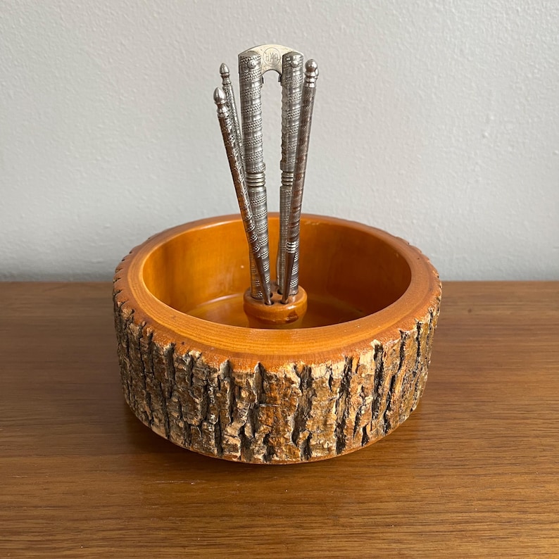 May include: A vintage nutcracker set featuring a wooden bowl with a bark-like exterior and a set of silver-toned nutcrackers. The bowl has a warm, orange-brown interior. The nutcrackers are arranged in the center of the bowl.