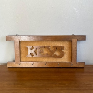Puede incluir: Portallaves de madera con la palabra "KEYS" tallada en el centro. El portallaves tiene un fondo de arpillera y varios ganchos para colgar llaves. El marco es de madera.