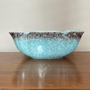 Puede incluir: Un macetero de cerámica turquesa con un esmalte marmoleado marrón y blanco. El macetero tiene un borde ondulado único y una forma ovalada ancha.