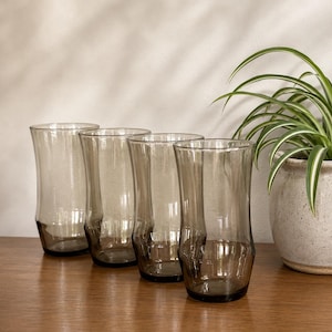 May include: Four tall, smoky grey drinking glasses with a curved design. The glasses are arranged on a wooden surface, with a potted plant in the background. The glasses have a unique shape, wider at the top and bottom.