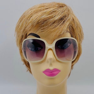 May include: Cream-colored, oversized sunglasses with a square frame and gradient lenses. The lenses transition from a dark purple at the top to a lighter shade at the bottom. The sunglasses are displayed on a mannequin head with blonde hair and pink lipstick.