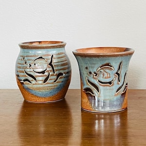 Puede incluir: Dos candelabros de cerámica con diseño de pez. Los soportes están esmaltados en tonos azules, marrones y naranjas. Los peces y las burbujas están recortados de la cerámica. Un soporte es redondo y el otro es un cilindro.