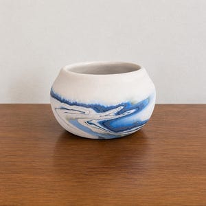 May include: A small, round ceramic bowl with a white exterior and a blue and white marbled design around the base. The bowl sits on a wooden surface. The interior of the bowl is white.