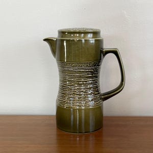 Może przedstawiać: Zielony dzbanek ceramiczny z fakturowanym wzorem i pokrywką. Dzbanek ma szeroką podstawę i wąską szyjkę.