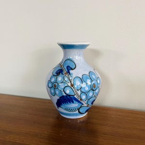 Peut inclure: Petit vase en céramique bleu clair avec un bord évasé et un corps arrondi. Le vase est décoré d'un motif floral dans les tons de bleu et de marron. Les fleurs sont stylisées et disposées en motif répétitif.