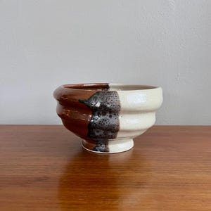 May include: A handcrafted ceramic bowl with a distinctive design. The bowl is split into two halves: one side is a deep brown, the other a creamy white. A textured, speckled black and grey section divides the colours. The bowl is displayed on a wooden surface.