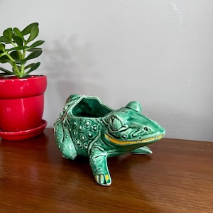 May include: A green ceramic frog planter with a textured surface and yellow accents. The frog has a wide open mouth and is sitting on a wooden surface. A red pot with a green plant is in the background.