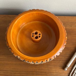 May include: A round, wooden bowl with a light brown finish. The bowl has a central, cylindrical holder with multiple holes. The rim of the bowl has a textured, bark-like edge. The bowl is likely for holding pencils or other small items.