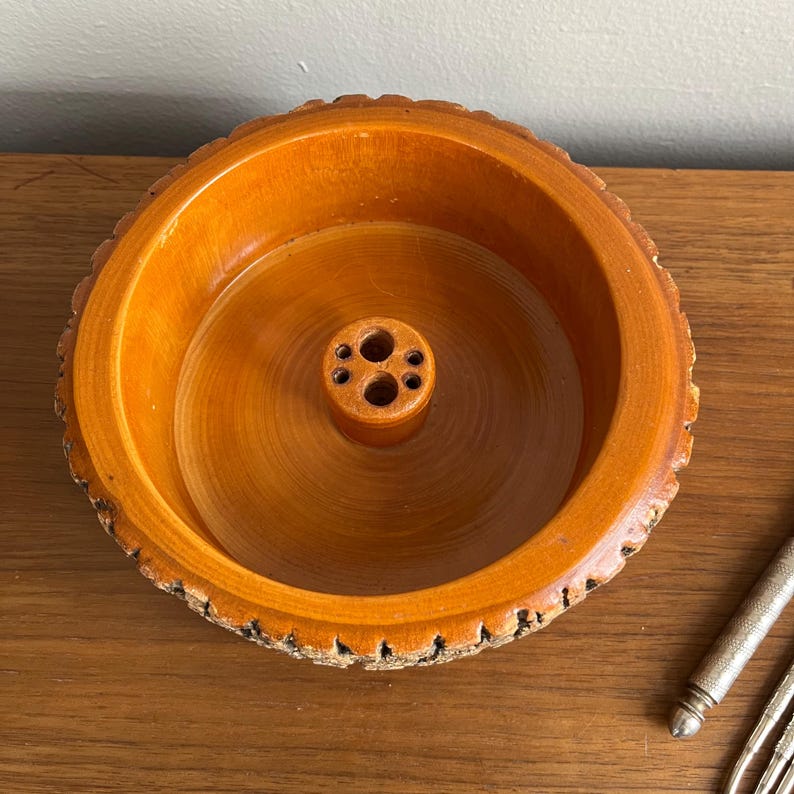 May include: A round, wooden bowl with a light brown finish. The bowl has a central, cylindrical holder with multiple holes. The rim of the bowl has a textured, bark-like edge. The bowl is likely for holding pencils or other small items.