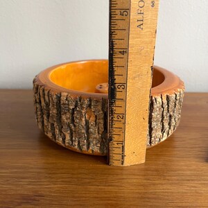 May include: A round, wooden bowl with a bark-like exterior and an orange interior. The bowl is approximately 5 inches in diameter and is being measured with a wooden ruler. The bowl is sitting on a wooden surface.