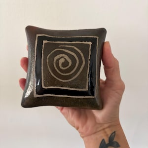 May include: A square ceramic dish with a dark brown, textured finish. The centre features a black square border surrounding a light-coloured, hand-painted spiral design. The edges of the dish are slightly curved, creating a soft, rounded square shape.