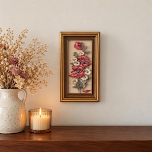 Puede incluir: Una pieza de arte mural rectangular en un marco dorado oscuro que presenta un arreglo floral tridimensional de amapolas rojas y pequeñas margaritas blancas sobre un fondo crema, junto a un jarrón de cerámica y una vela.