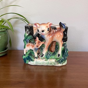 Puede incluir: Una maceta de cerámica con una cierva y su cervatillo. Los ciervos son de color marrón claro con manchas blancas, sobre un fondo de follaje verde y tocones de árboles negros. La maceta tiene forma rectangular y probablemente es vintage.