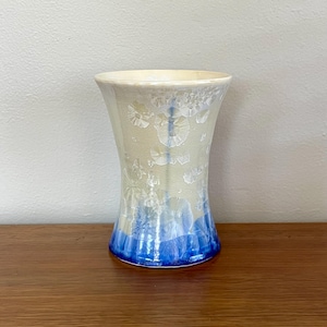 May include: A handmade ceramic vase with a flared design. The vase has a cream-coloured body with blue base and accents. The surface has a distinctive crystalline texture, adding visual interest. Suitable for displaying flowers or as a decorative item. Measures approximately 15 cm in height.