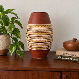May include: A barrel-shaped ceramic vase with a smooth terracotta-coloured top and bottom. The centre features horizontal bands of cream, tan, and dark brown with intricate embossed geometric patterns, including cross-hatching and dotted lines.