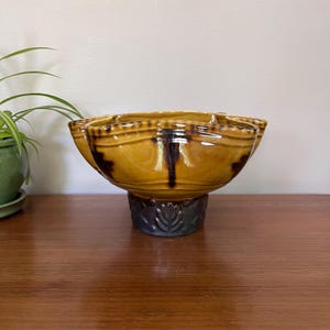 Può includere: Una ciotola decorativa in ceramica con una forma e un design unici. La ciotola è di colore giallo caldo con accenti marrone scuro e poggia su un piedistallo grigio scuro. Il piedistallo ha un motivo floreale. La ciotola è vuota.