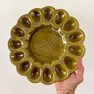 Puede incluir: Un plato para huevos redondo de cerámica verde oliva con borde festoneado y doce hendiduras individuales en forma de huevo. El centro del plato muestra una ilustración detallada en relieve de un gallo junto a una pequeña flor.