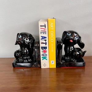 May include: A pair of black ceramic dog bookends with red detailing. Each bookend has a glossy black dog figurine with blue eyes and a red tongue. Two books are held between them, one titled "THE ART BOOK" and the other "ART DECO".