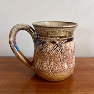 Puede incluir: Taza de cerámica hecha a mano con una superficie texturizada y un asa curva. La taza presenta una combinación de colores marrón y beige con detalles azules en el asa. La taza tiene un diseño tallado único en la parte delantera. Para café, té u otras bebidas.