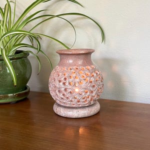 Könnte beinhalten: Ein rosa, geschnitzter Duftdiffusor aus Stein mit floralem Design. Der Diffusor hat einen runden Sockel und einen vasenartigen Aufsatz. Die filigranen Schnitzereien lassen Licht durchscheinen und erzeugen ein warmes Leuchten. Eine grüne Topfpflanze befindet sich im Hintergrund.