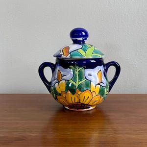 May include: A hand-painted ceramic sugar bowl with a lid. The bowl is decorated with a floral design in yellow, blue, green, and orange. The lid has a blue knob. The sugar bowl has two handles.