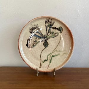 Könnte beinhalten: Ein Keramikteller mit handgemaltem Schmetterlingsdesign. Der Schmetterling hat blaue, grüne und schwarze Details. Der Teller hat einen beigen Hintergrund mit braunem Rand und steht auf einem dekorativen Metallständer.