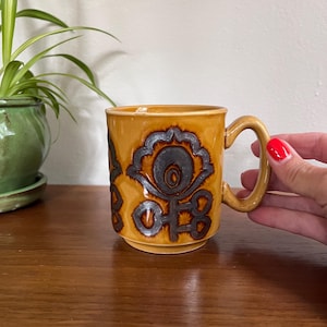 May include: A tan ceramic mug with dark brown floral patterns. The mug has a curved handle. The mug is held by a person. The mug is on a wooden surface.