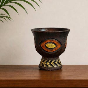 May include: A dark brown, vintage ceramic vessel with an eye-shaped design in orange and yellow. The vessel has a textured surface and a pedestal base with a repeating arrow pattern in gold and yellow. It sits on a wooden surface.