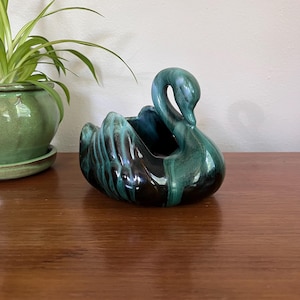 May include: A ceramic swan-shaped planter featuring a glossy glaze with blended shades of deep forest green, teal, and black. The swan has a curved neck and detailed wing textures, with an open basin in the center for holding small items or plants.