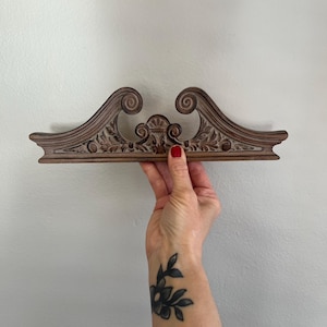 May include: A decorative wooden trim with intricate carvings, featuring a scroll design and floral motifs. The trim is painted in a light brown color with a distressed finish.