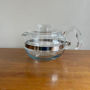 May include: A clear glass teapot with a lid and handle. The teapot has a silver band around the middle. The teapot is sitting on a wooden surface.