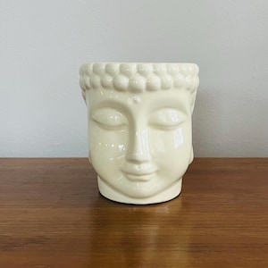 Puede incluir: Un macetero de cerámica blanca con forma de rostro de Buda con los ojos cerrados. El macetero tiene un acabado liso y brillante.