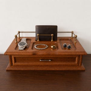 May include: A wooden valet tray featuring a single drawer with a metal handle, a brass-toned railing, and three recessed compartments. The tray holds a silver-tone wristwatch, a silver-tone chain bracelet, a gold-tone ring, two cufflinks, and a dark brown wallet.