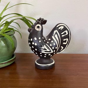 Puede incluir: Figura de gallo de cerámica negra con lunares blancos y detalles decorativos blancos. El gallo tiene una cresta y plumas de cola detalladas. La figura se exhibe sobre una superficie de madera, con una planta en maceta en el fondo.