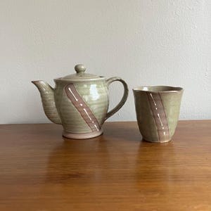 May include: A ceramic tea set featuring a teapot with a lid and a matching cup. The set is glazed in a muted green with a diagonal brown stripe accented with white dashes. The teapot has a curved spout and handle.