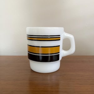 May include: White ceramic mug with a handle, featuring horizontal stripes in black, gold, and white. The mug has a stacked design and a simple, rounded handle. The mug is sitting on a wooden surface.
