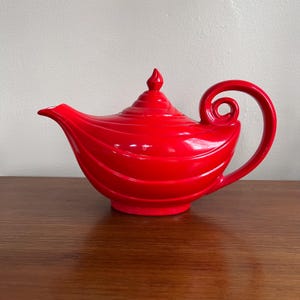 May include: A bright red ceramic teapot with a curved spout and a looped handle. The teapot features a ridged design and a small finial on the lid. The teapot is a decorative item.