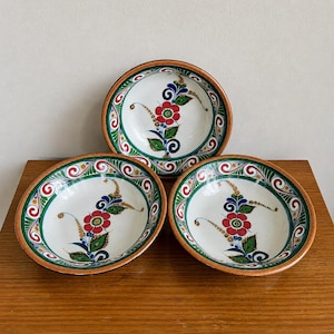 May include: Three ceramic bowls with a floral design. Each bowl has a white interior with a red flower, green leaves, and blue accents. The rims are decorated with green, red, and gold patterns, and have a brown border.