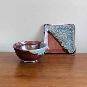 Puede incluir: Un juego de cuenco y plato cuadrado de cerámica hecho a mano. El cuenco es de color rojo intenso con detalles azules y blancos. El plato presenta un diseño rojo y azul con detalles marrones y blancos. El conjunto está sobre una superficie de madera.