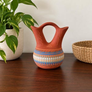 May include: A terracotta-coloured ceramic vessel featuring a double-spout design connected by a central arched handle. The lower body is decorated with horizontal bands of geometric patterns in shades of blue, white, and orange-red.