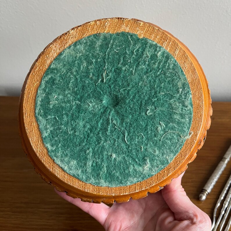 May include: A round, wooden bowl with a green felt interior. The bowl's exterior is a light brown color with a textured, ridged edge. The bowl is being held by a person's hand.