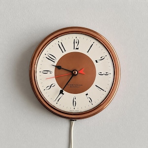 Könnte beinhalten: Eine runde Wanduhr in Kupferfarbe mit cremefarbenem Zifferblatt. Sie hat schwarze Ziffern, schwarze Zeiger und einen roten Sekundenzeiger. Die Mitte ist dunkler kupferfarben. Der Text "Westclox electric" ist auf dem Zifferblatt aufgedruckt.